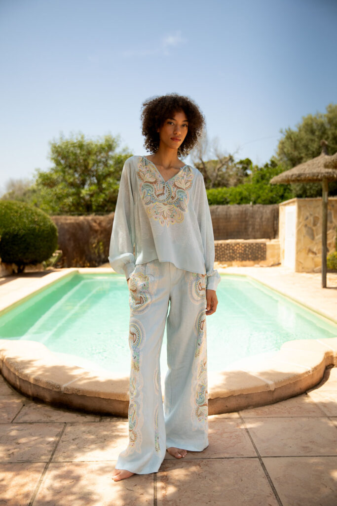 Imagebild: Model steht vor Pool und trägt EMBELLISHED LACE Seidenbluse und Leinenhose.