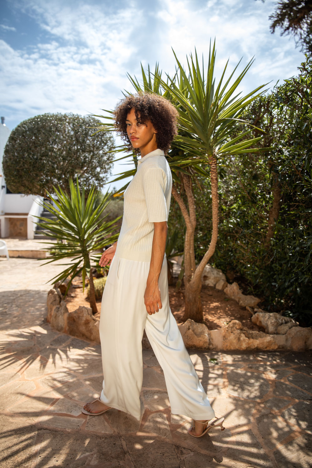 Imagebild: Model steht vor Palmen und trägt SOLID KNIT Kaschmir Poloshirt und Seidenhose in weiß.