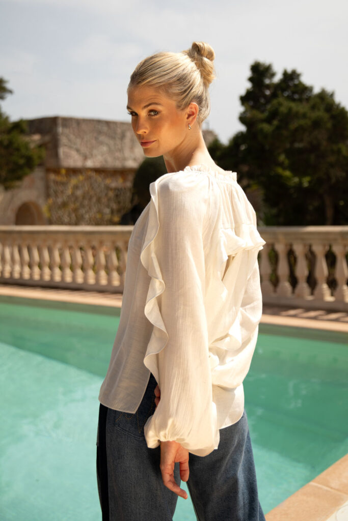 Imagebild: Model steht vor Pool vor Finka und trägt FRILL Bluse in weiß mit Jeans.
