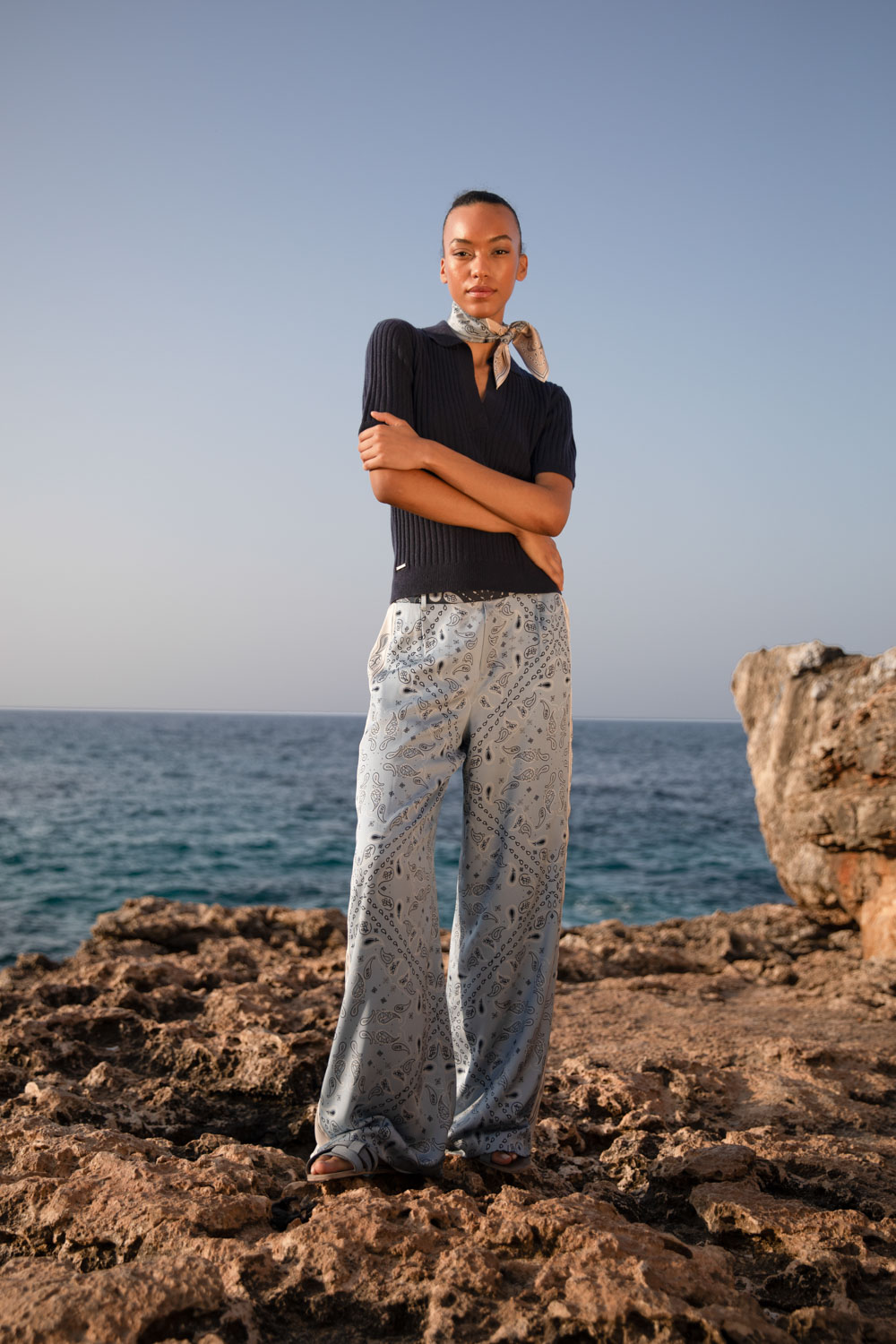 Imagebild: Model steht auf Küstenklippen und trägt BANDANA PATCH Seidentuch, SOLID KNIT Kaschmir Poloshirt in dunkelblau und BANDANA PATCH Seidenhose.