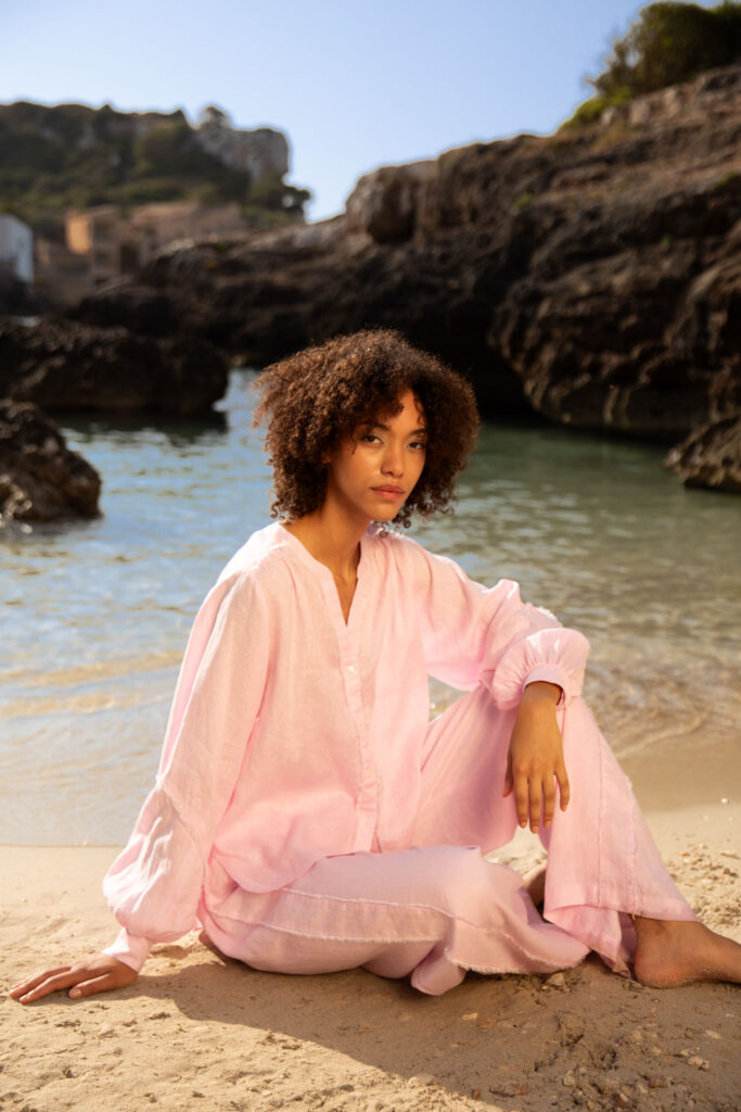 Imagebild: Model sitzt im Strand vor dem Meer und trägt SOLID LINEN Bluse und Hose in rosa.
