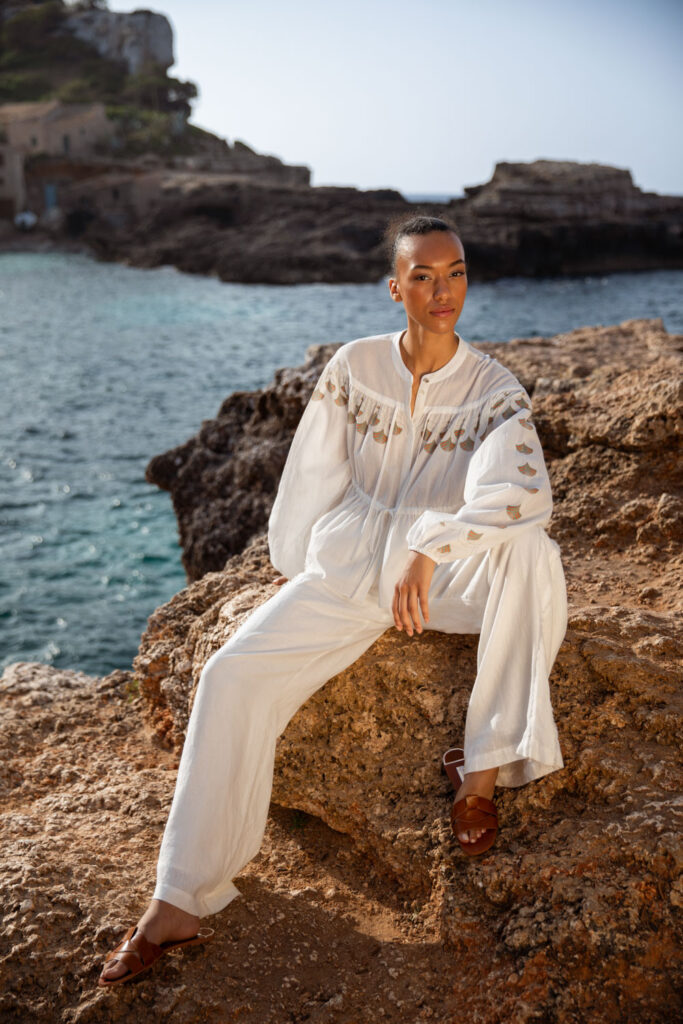Imagebild: Model sitzt auf Klippen, das Meer im Hintergrund und trägt ABANICO Baumwollbluse in weiß mit SOLID LINEN Leinenhose in weiß.