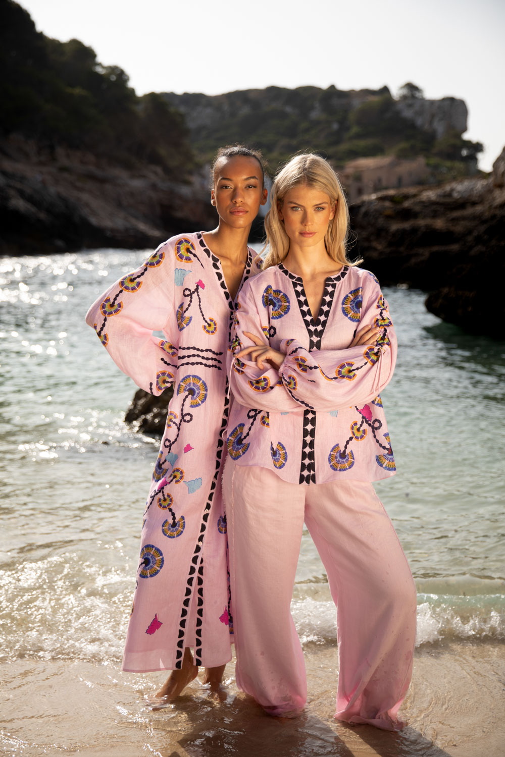 Imagebild: Models stehen im Sand vor einer Meeresbucht. Ein Modell trägt das FOLDED FLOWERS Leinenkleid. Das andere Model trägt FOLDED FLOWERS Leinenbluse und SOLID LINEN Leinenhose in rosa.
