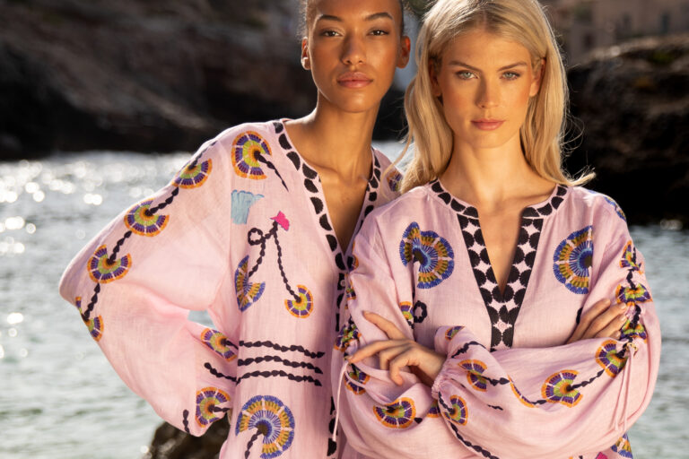 Imagebild: Zwei Models stehen im Sand vor einer Bucht auf Mallorca. Sie tragen unseren FOLDED FLOWERS Print: ein graphischer floraler Print auf rosafarbenem Stoff aus Leinen.