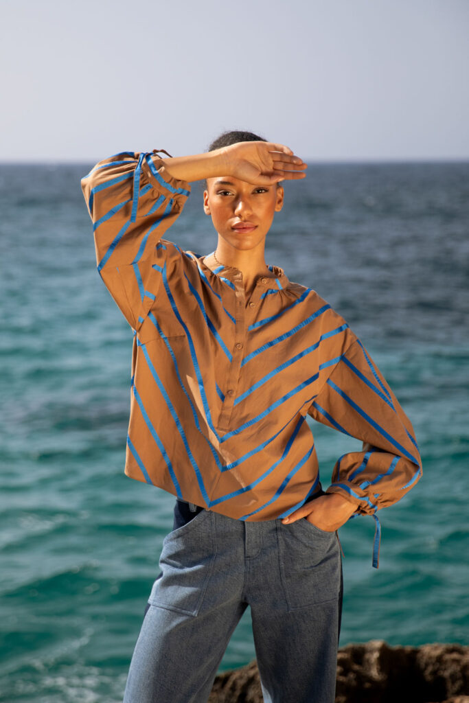 Imagebild: Model steht auf Küstenklippen vor dem Meer und trägt die ROYAL STRIPE Baumwollbluse.